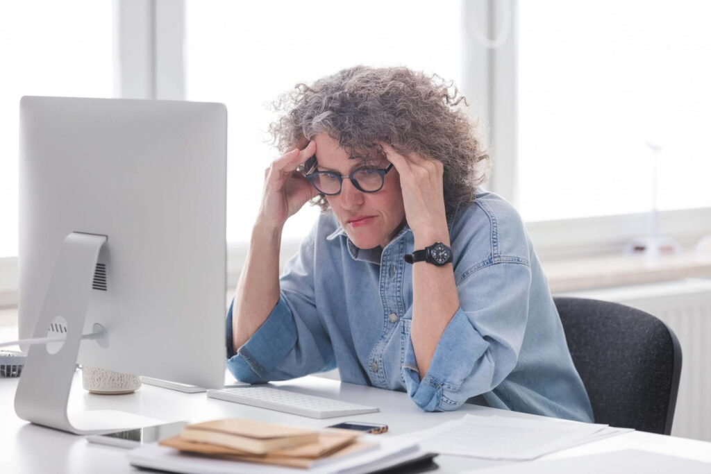 Una mujer sentada en un escritorio frente a una computadora, con las manos a los lados de la cabeza y parece estresada.