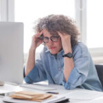 Una mujer sentada en un escritorio frente a una computadora, con las manos a los lados de la cabeza y parece estresada.